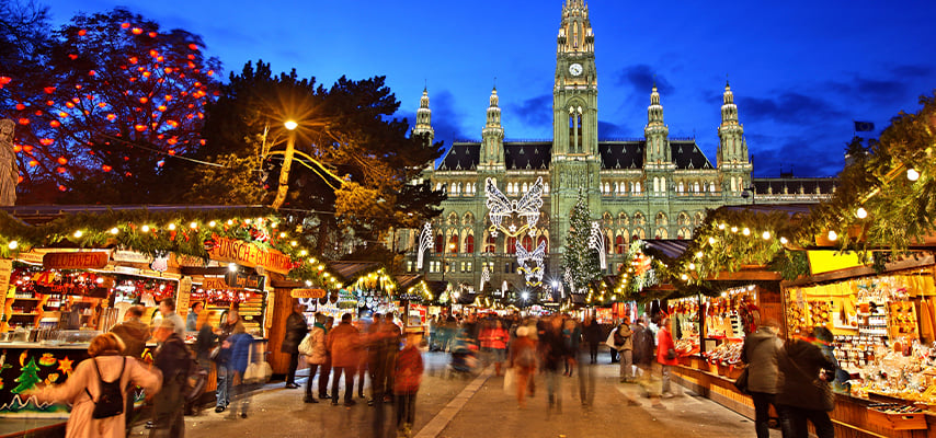 Busy Christmas market with wooden stalls fills square in front of illuminated Gothic city hall in Vienna Austria.