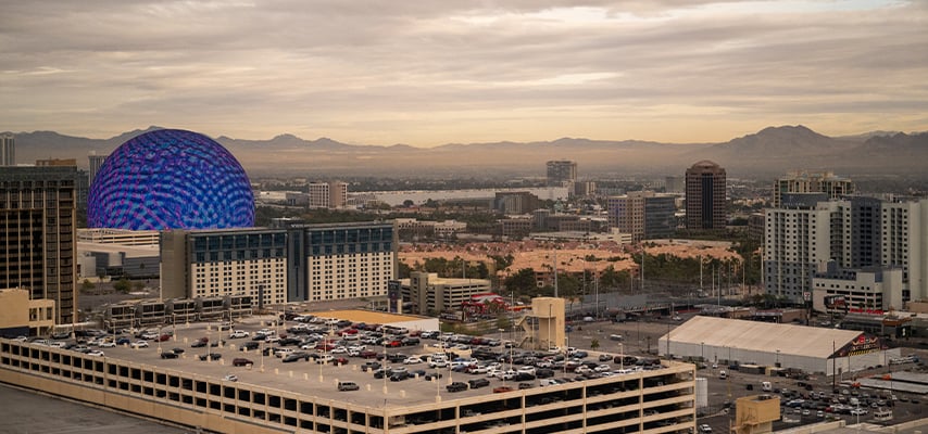 The Sphere venue glows blue and purple against a cloudy dusk sky in an elevated view of the Las Vegas east side and surrounding desert.