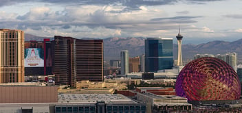 Aerial view of the Las Vegas Strip skyline with the Sphere venue displaying a colorful pattern, the Stratosphere tower, and desert mountains in the background.