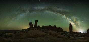 The Milky Way arches across a star-filled green sky above silhouetted sandstone rock formations in Utah's desert landscape at night.