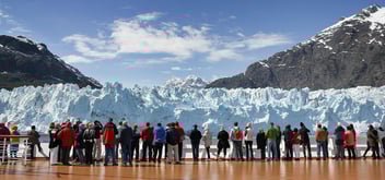 Passengers on the deck of a cruise ship looking at a glacier and the mountainous landscape of Alaska.