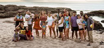 Extended family of multiple generations poses together on sandy beach with rocky coastline and ocean backdrop.